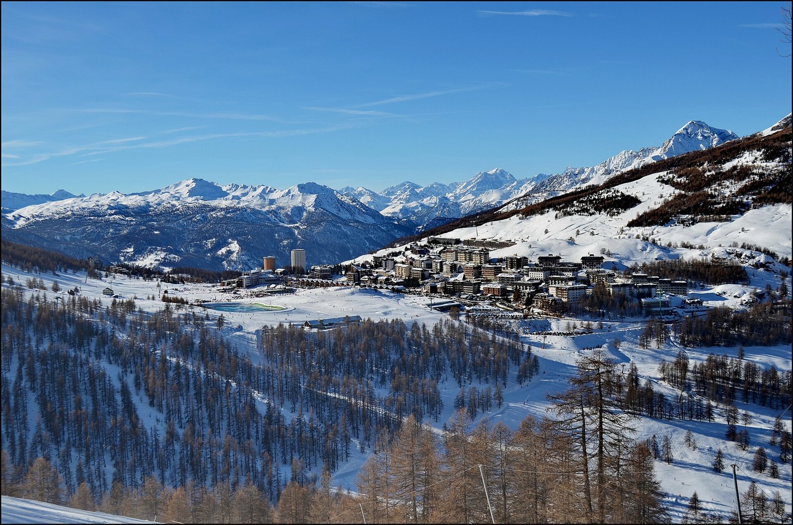 Sestriere innevata vista dall'alto, con il paese, le piste, i boschi di larici e le montagne sullo sfondo